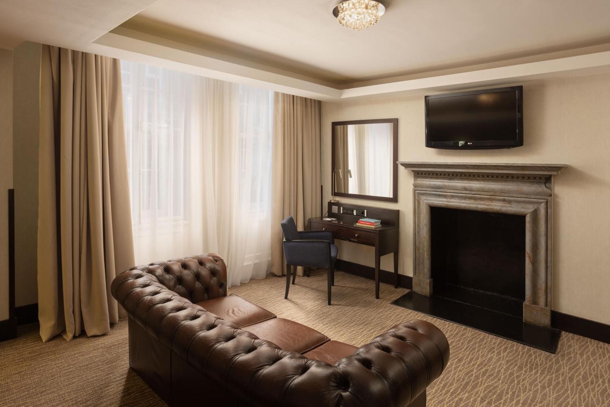 Hotel Montcalm Brewery Townhouse Londres Exterior foto The photo shows a stylish interior of a room, likely a living area or a hotel suite. It features a dark leather chesterfield sofa with tufted upholstery. Opposite the sofa is a small wooden desk with a chair, positioned near a wall-mounted flat-scree