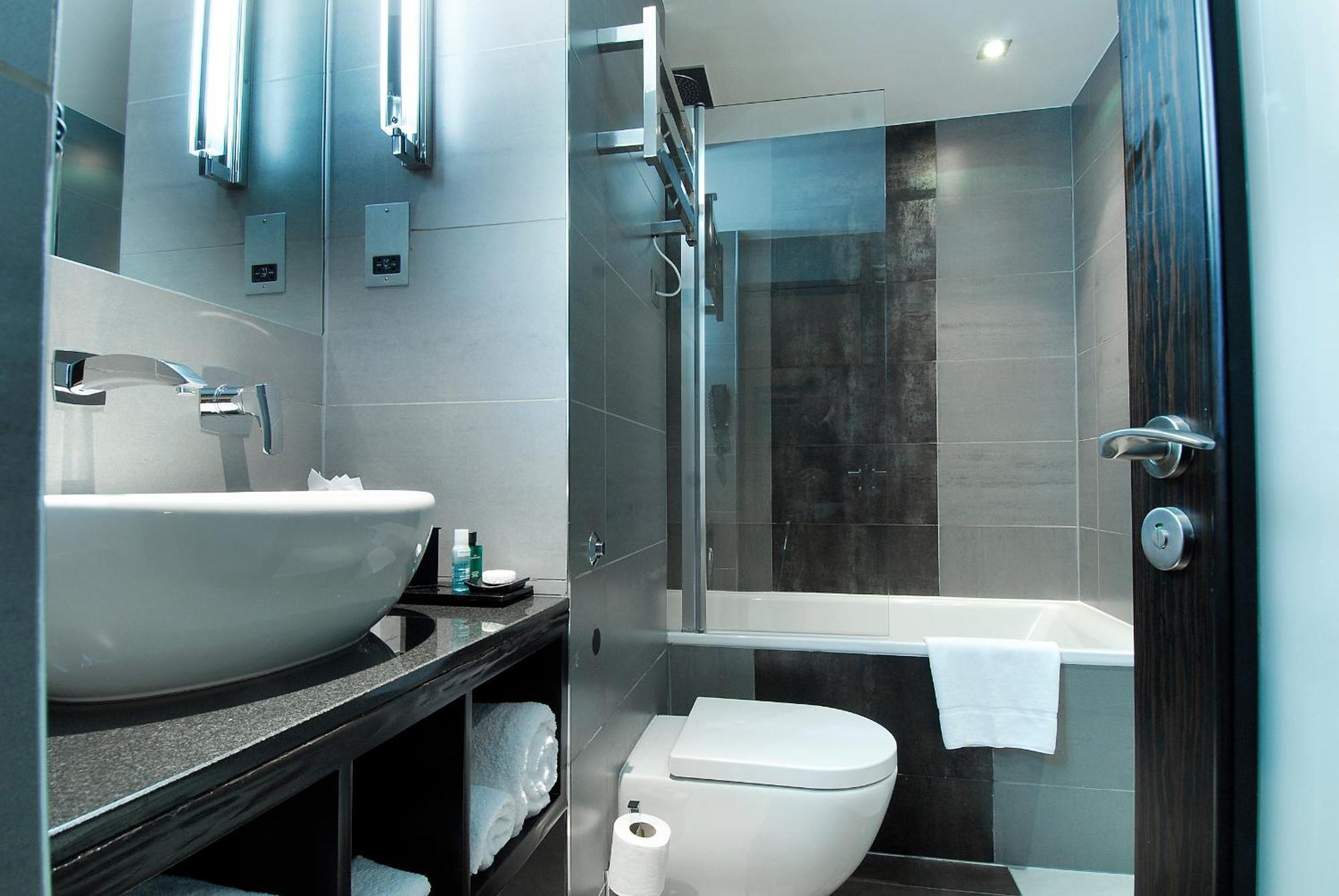 Hotel Montcalm Brewery Townhouse Londres Exterior foto The photo shows a modern bathroom with sleek design elements. It features a curved white sink on a dark countertop, with a mirror above that is illuminated by stylish lighting fixtures. Next to the sink, there's a toilet, and on the opposite side, a 