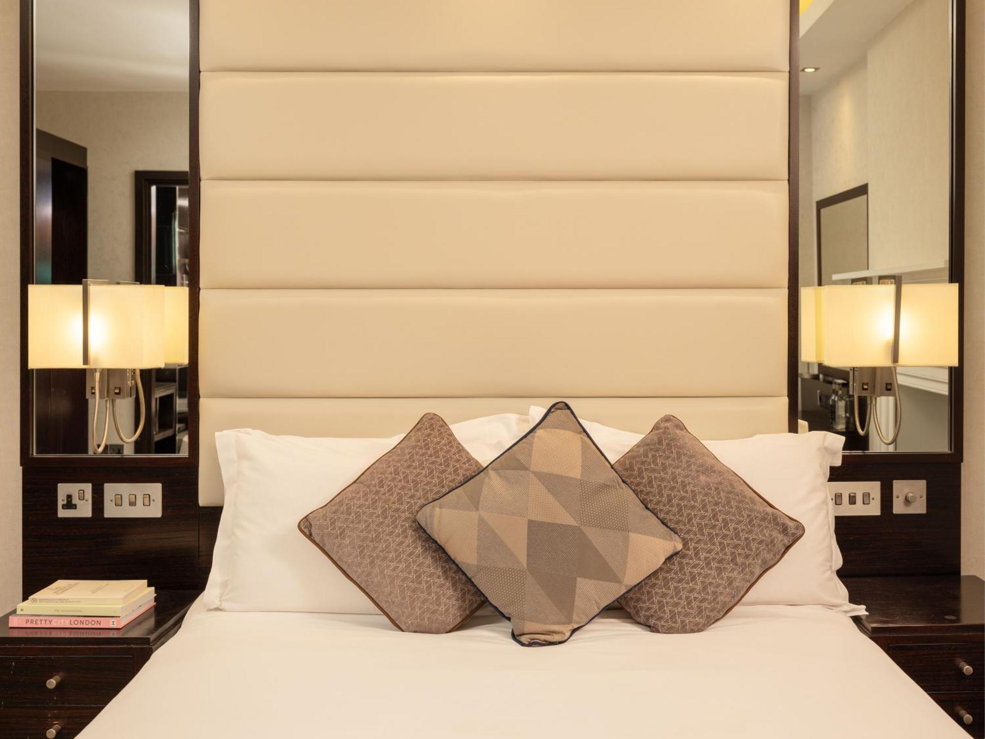 Hotel Montcalm Brewery Townhouse Londres Exterior foto The image shows a cozy bedroom setting. The bed features a headboard upholstered in a light beige fabric. It is adorned with three decorative pillows in varying patterns and colors, placed on white bedding. On either side of the bed, there are modern