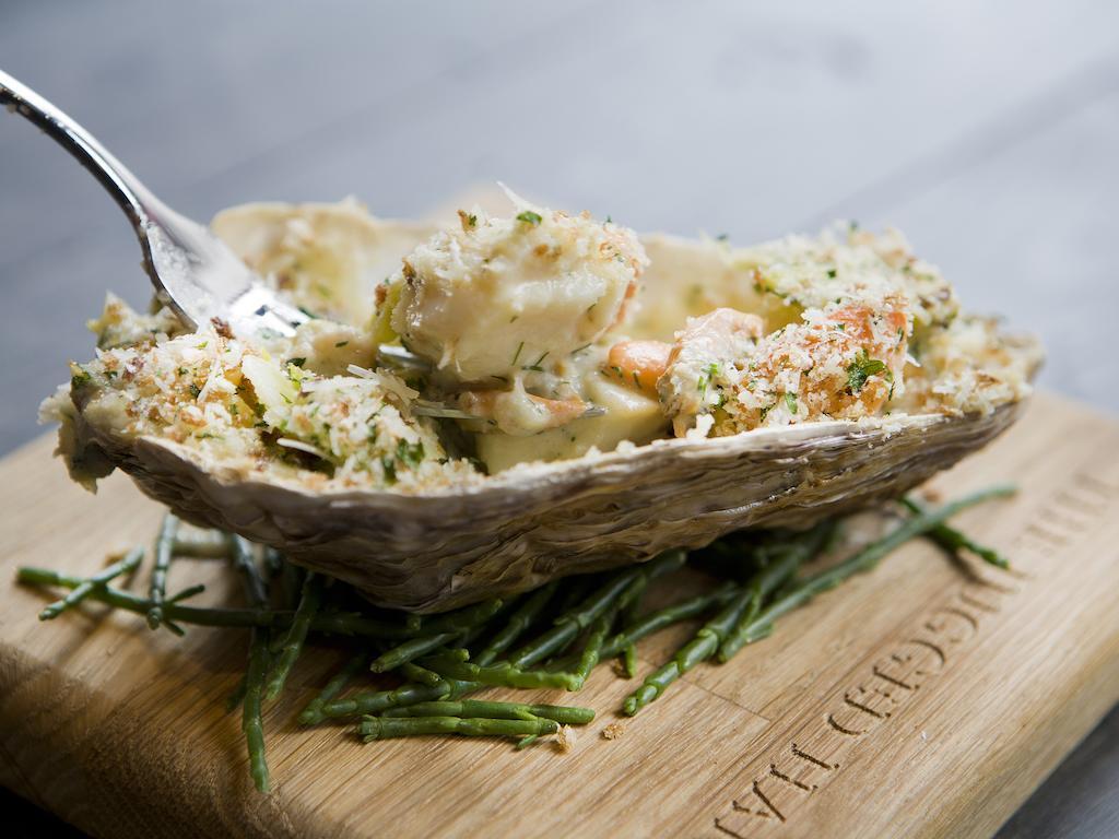 Hotel Montcalm Brewery Townhouse Londres Exterior foto The photo shows a large oyster shell filled with a mixture that appears to include seafood, herbs, and breadcrumbs. The dish is presented on a wooden board with some greenery, possibly a type of seaweed or garnishing. A fork is placed beside the oyst