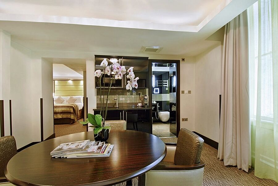 Hotel Montcalm Brewery Townhouse Londres Exterior foto The photo shows a stylish hotel room or suite. In the foreground, there is a round dining table with a few magazines on it. Surrounding the table are upholstered chairs. To the right, there are large windows with light-colored curtains, allowing natu